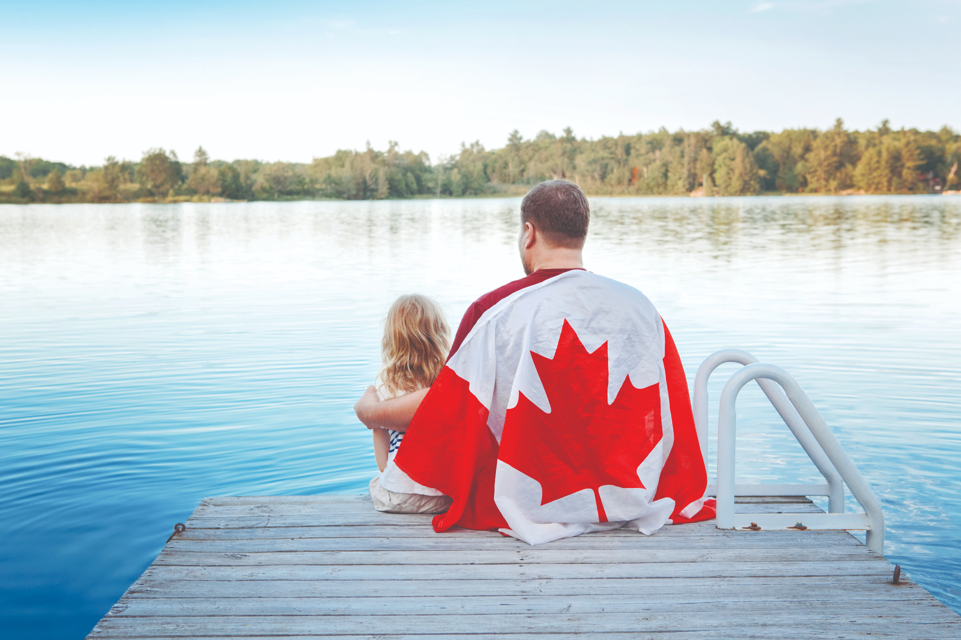 Father and child wrapped in a Canadian flag sitting on a lakeside dock